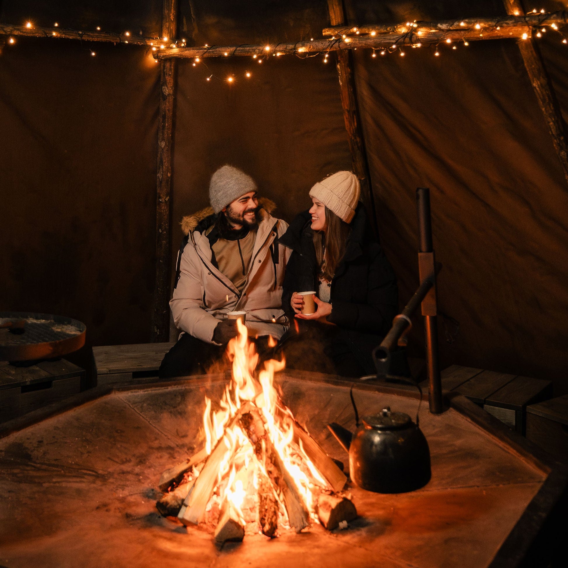 Two people sitting by a campfire inside a tent with string lights.