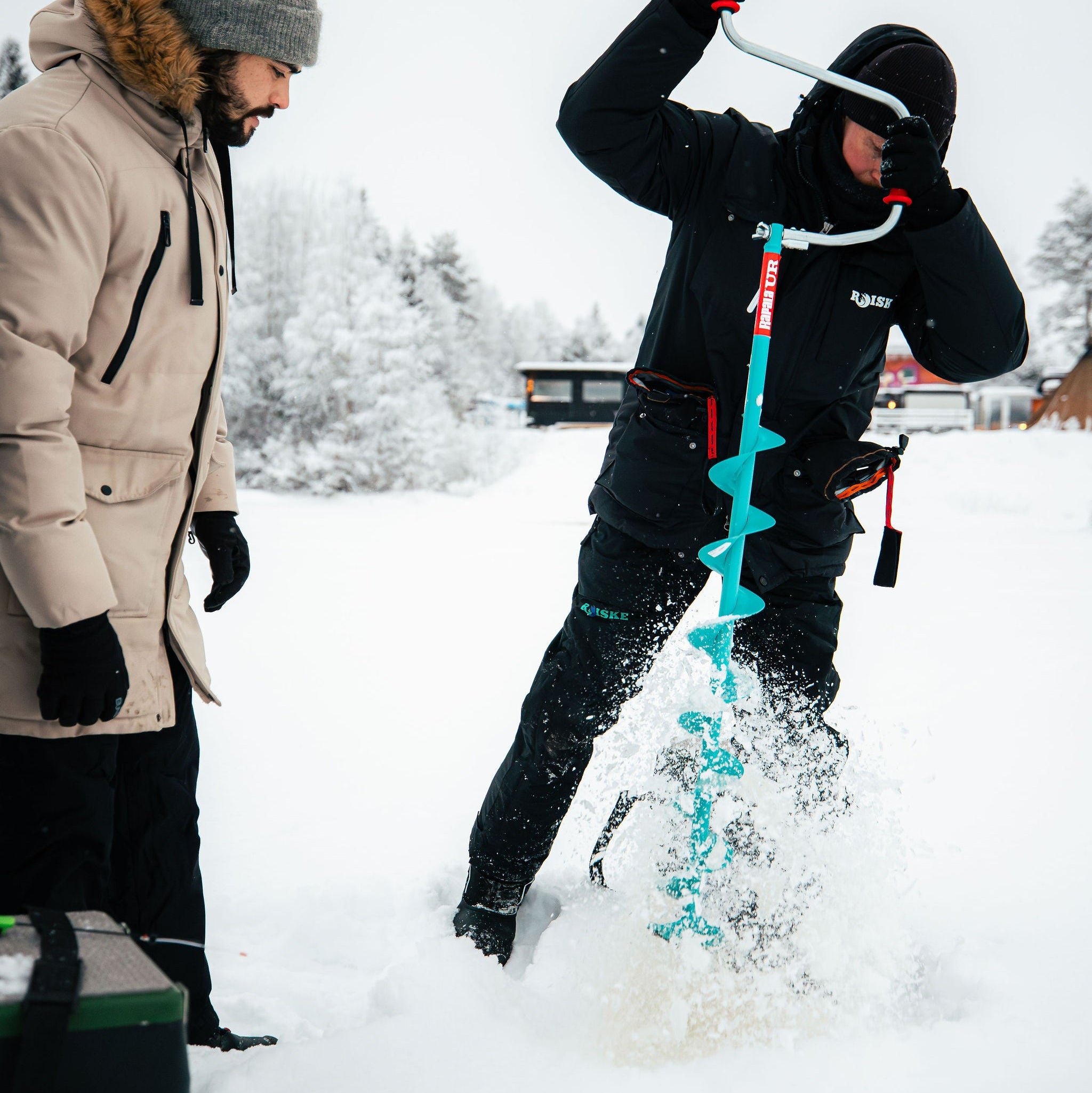 Person using an ice auger to drill a hole in the snow, with another person observing.