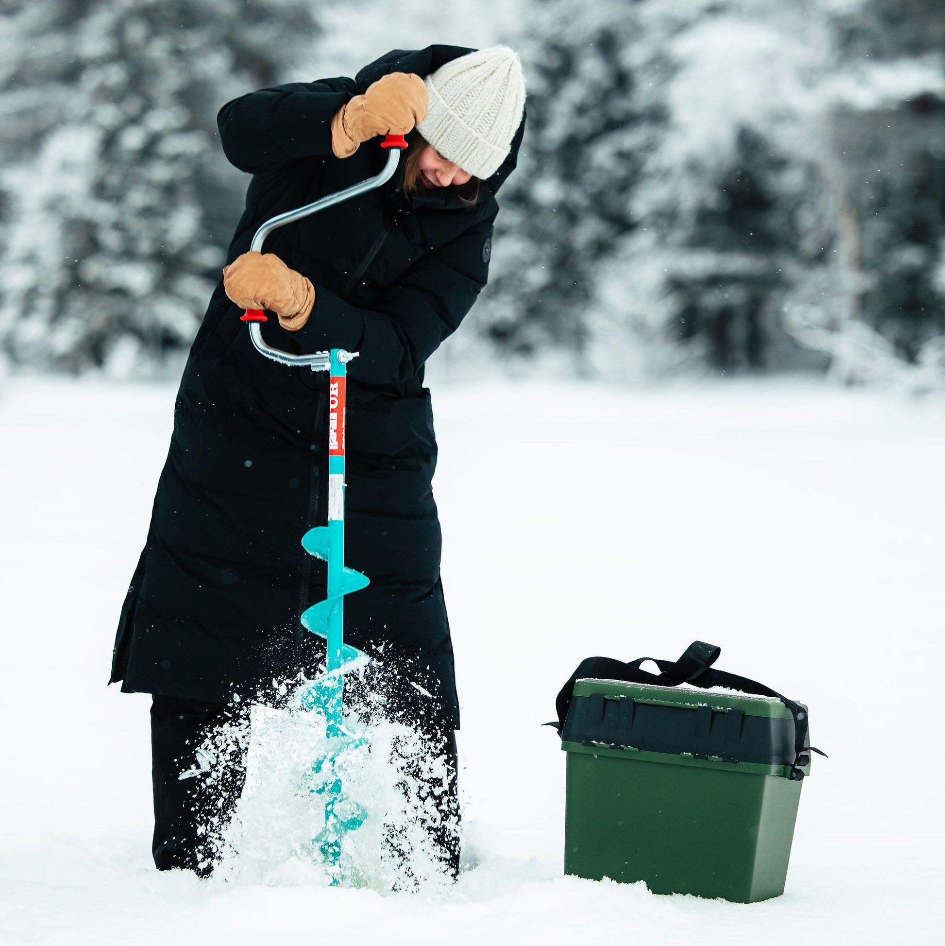 Person using an ice auger in the snow with a green box nearby