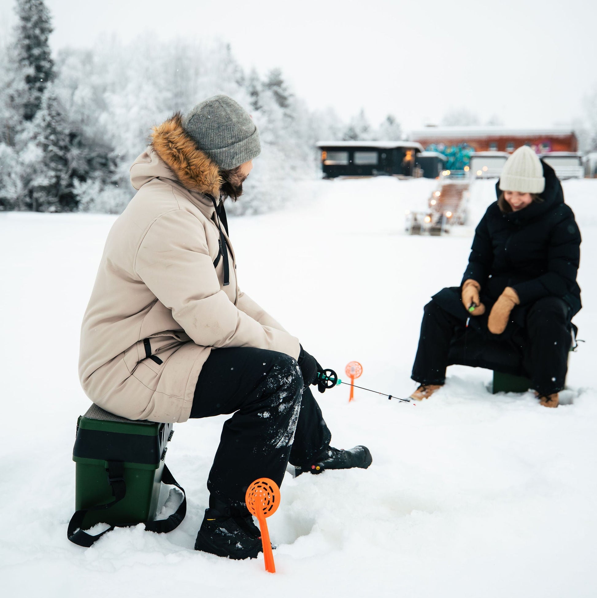 Two people ice fishing on a frozen lake with snow-covered trees and a building in the background.