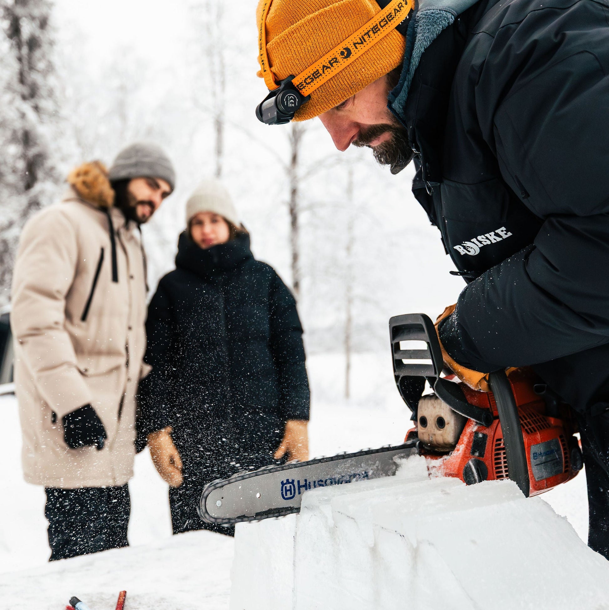Person using a chainsaw to cut ice in a snowy landscape with two observers.