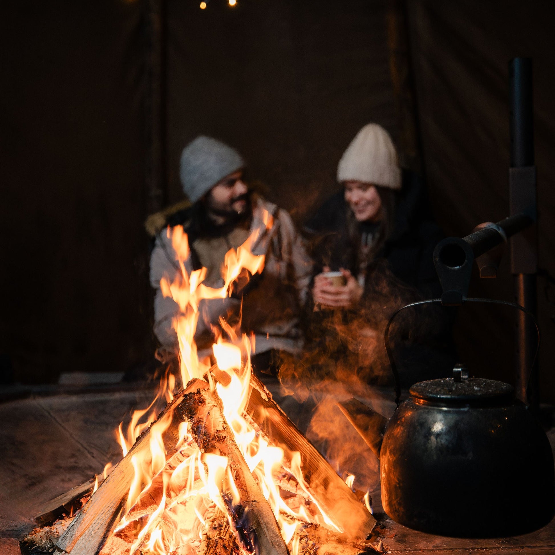 Two people sitting by a campfire inside a tent with string lights above.