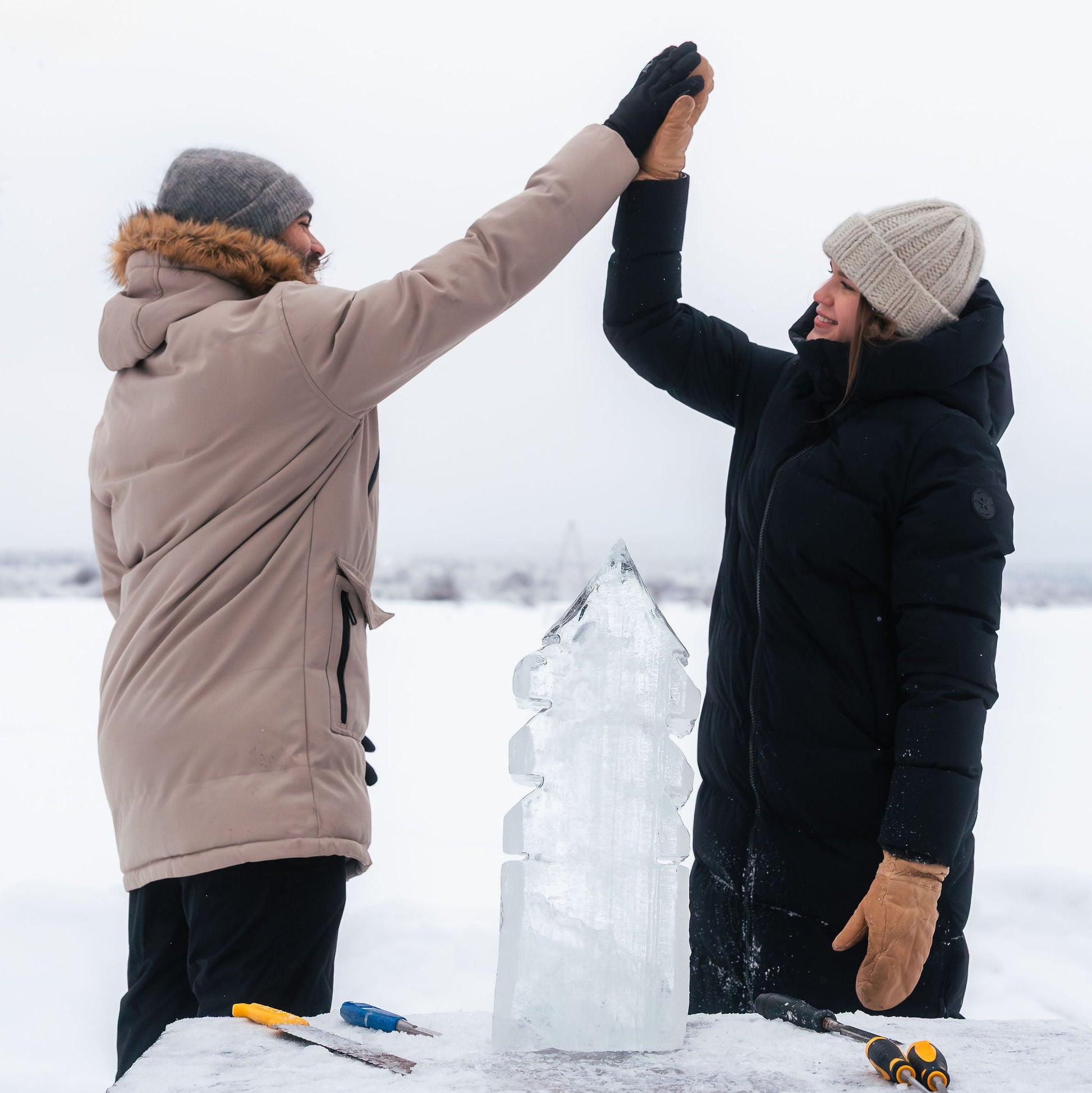 Two people in winter clothing working on an ice sculpture in a snowy landscape.