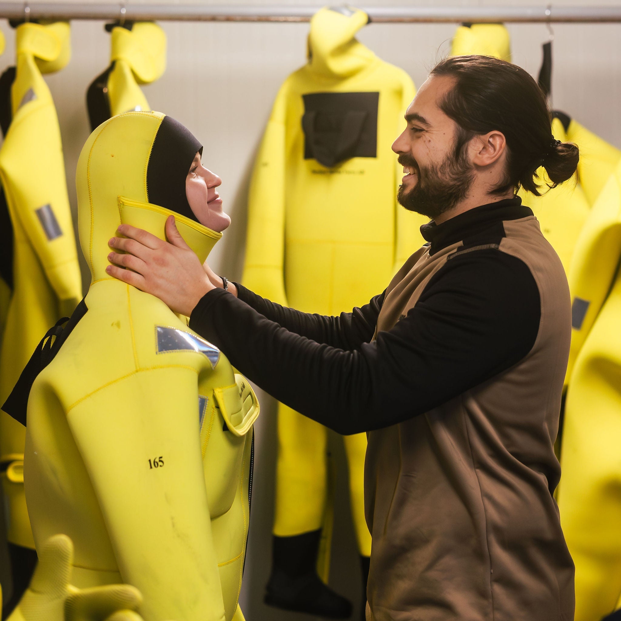 Two people in a room with yellow jackets, one person is adjusting the jacket of the other.