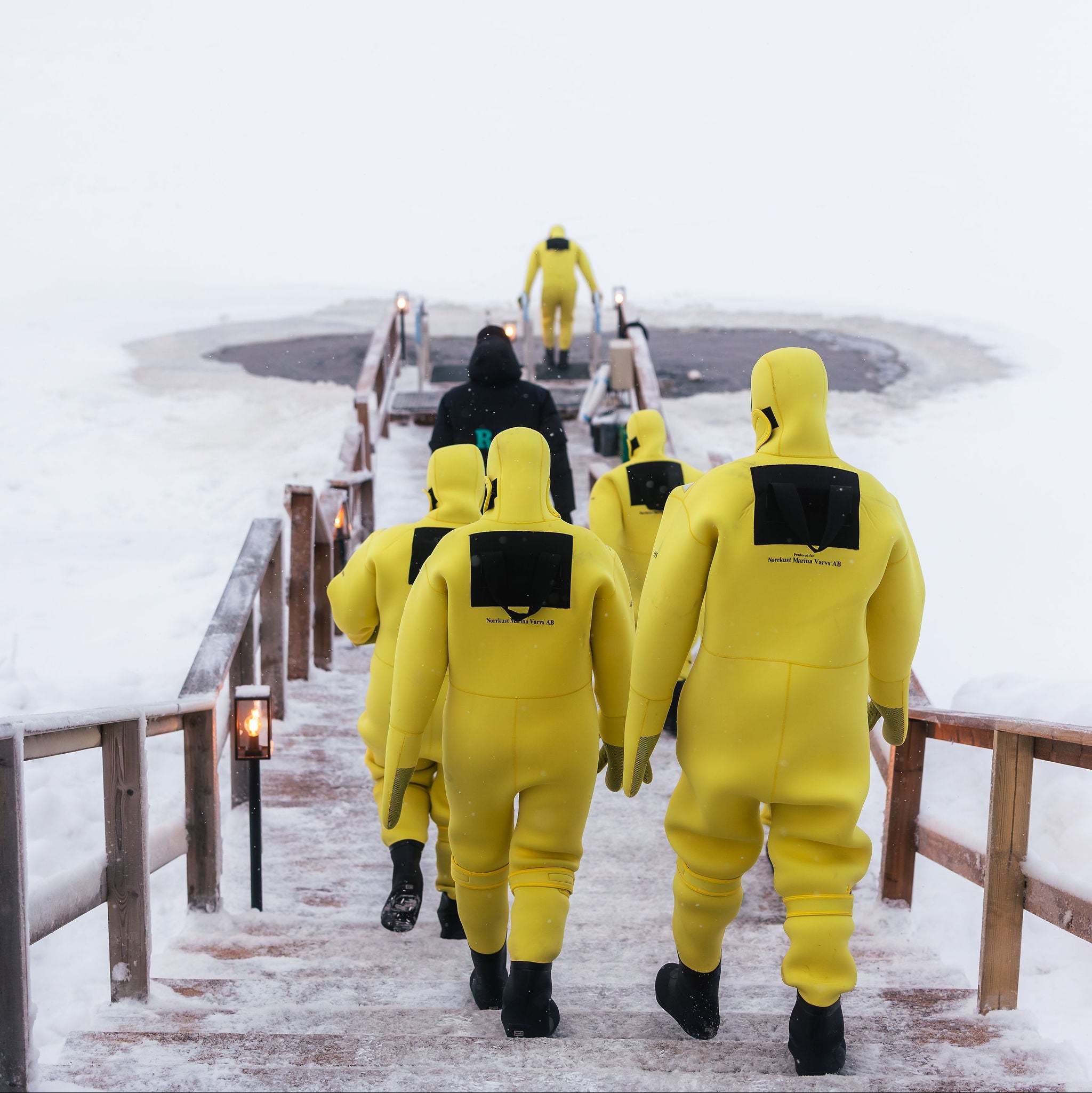 People in yellow suits walking on a snowy path with a frozen lake and pink sky in the background.