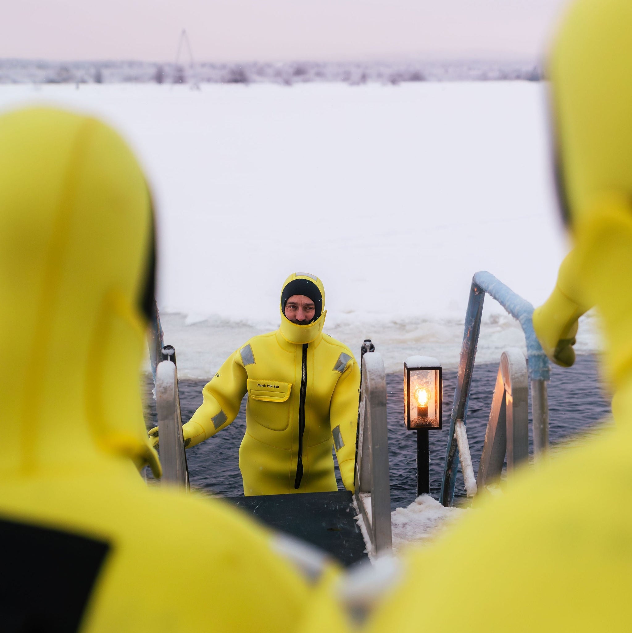 Person in a yellow raincoat standing on a snowy deck with a lantern in the background