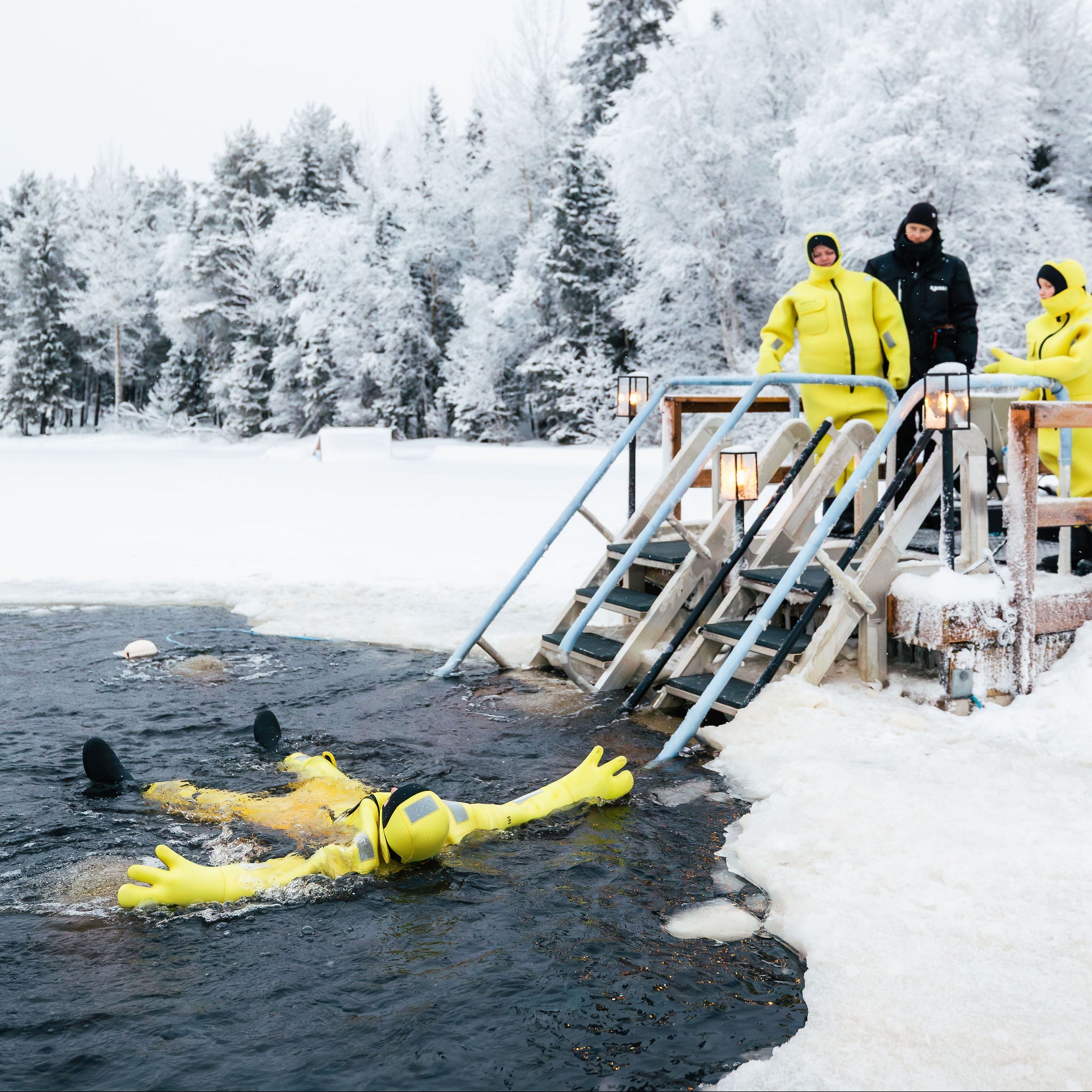 Person in a yellow wetsuit swimming in a frozen lake with others on a wooden platform in the background.