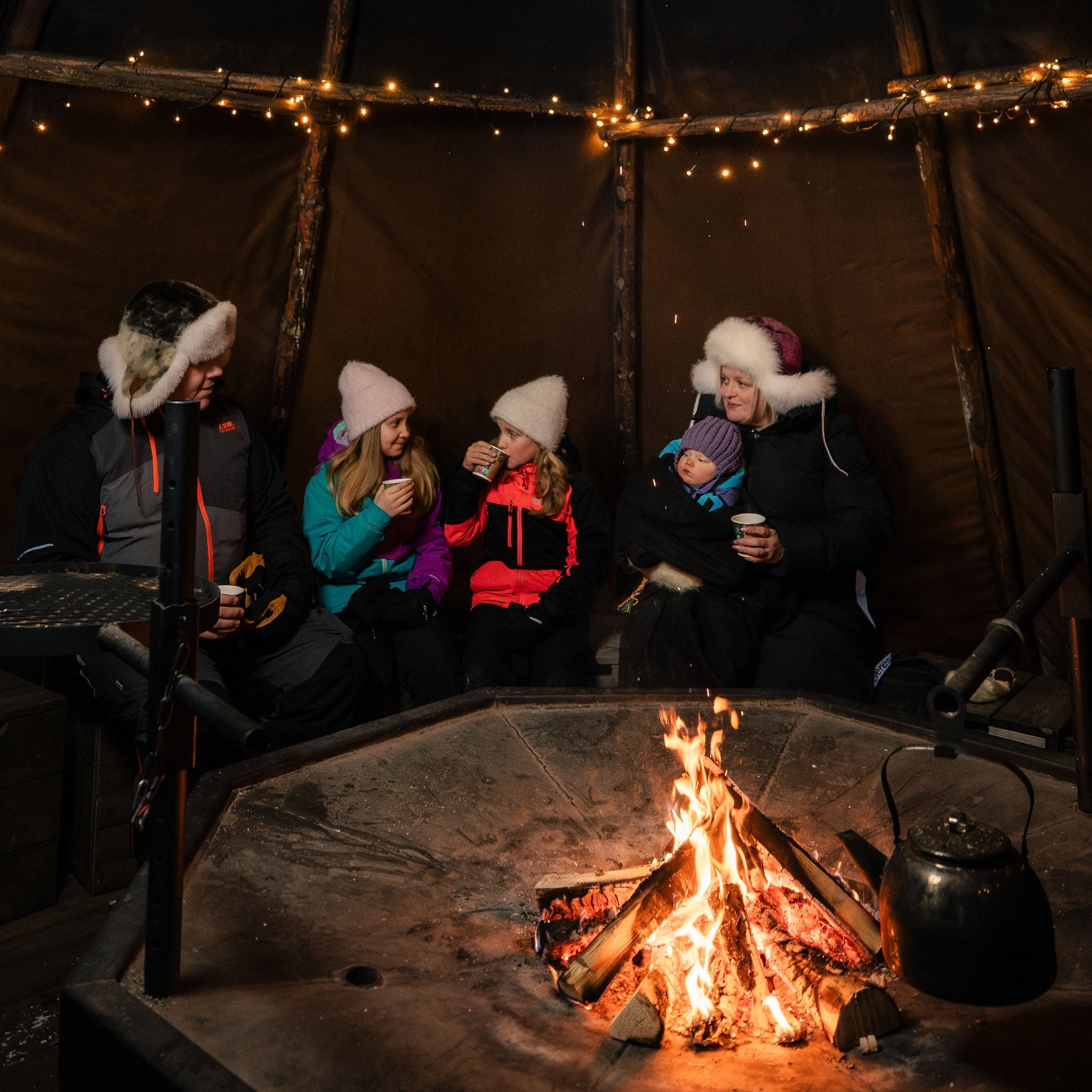 People sitting around a campfire inside a tent with string lights.