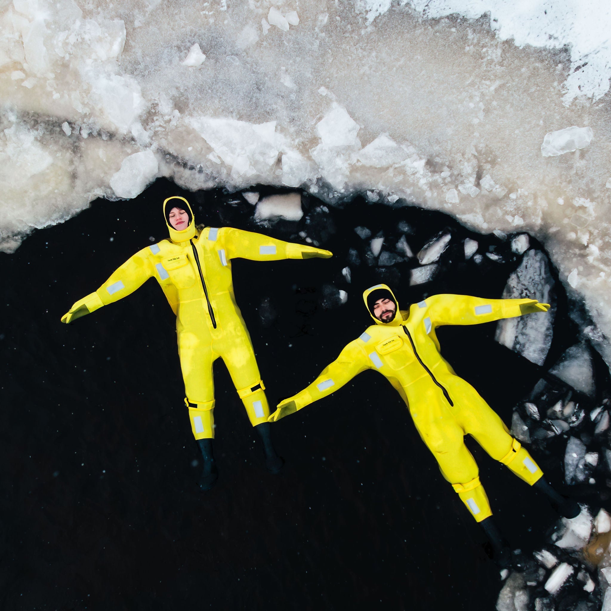 Two people in yellow rain suits lying on a frozen surface with ice cracks.
