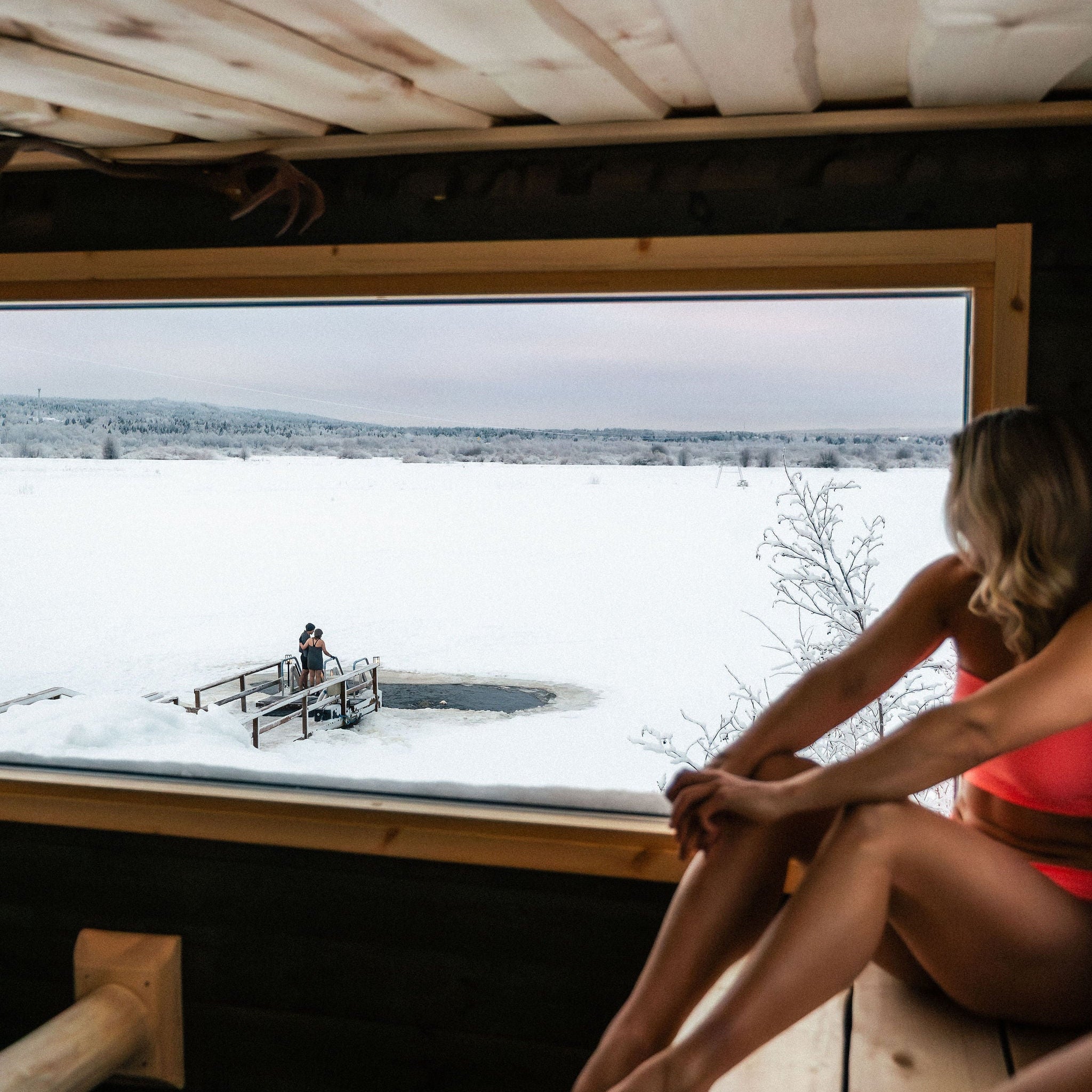 Person sitting in a sauna looking out at a snowy landscape with a small dock.