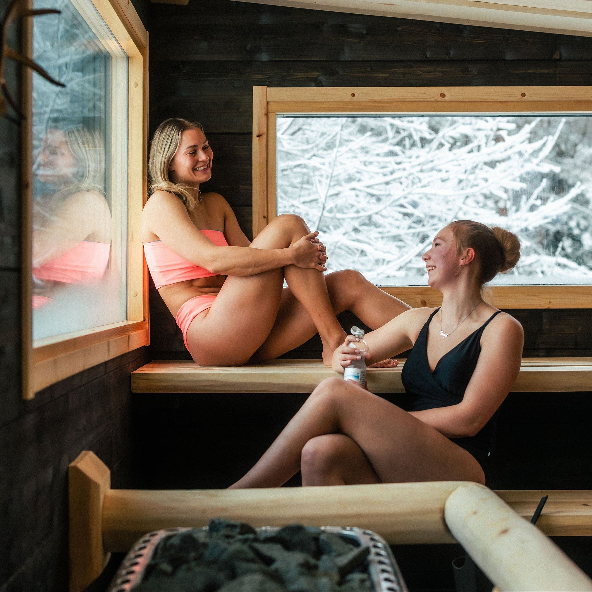 Two women sitting in a wooden sauna with a snowy landscape outside.