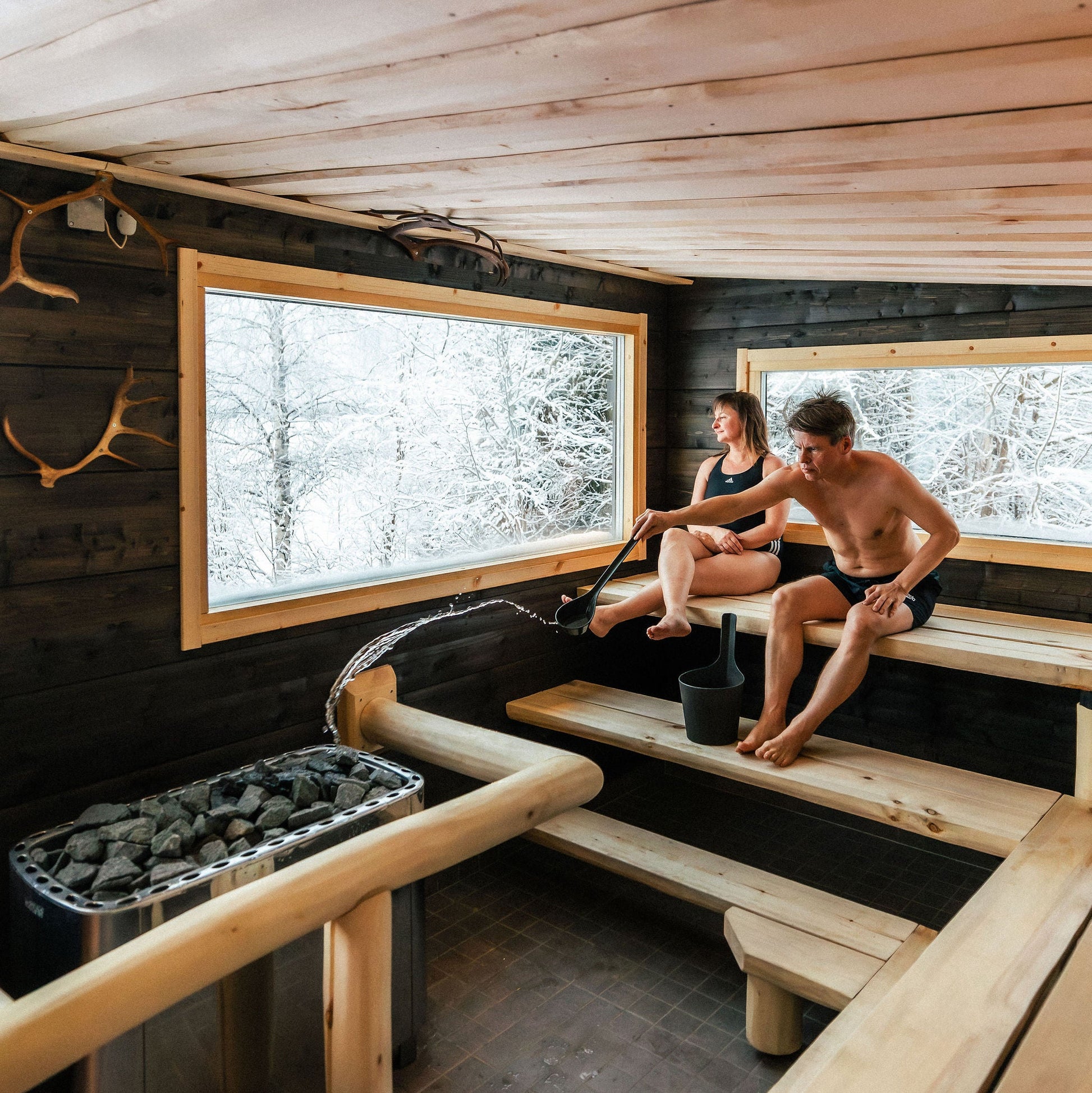 Two people sitting in a wooden sauna with large windows showing a snowy landscape.
