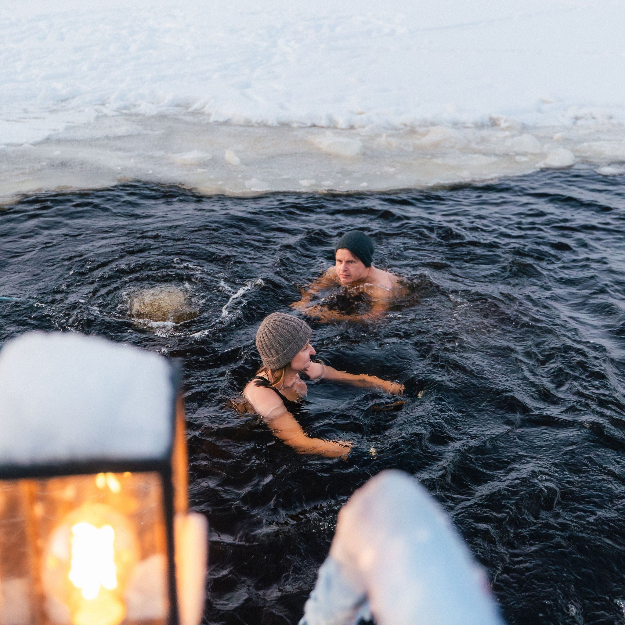 Two people in a icy water with a firelit tent nearby