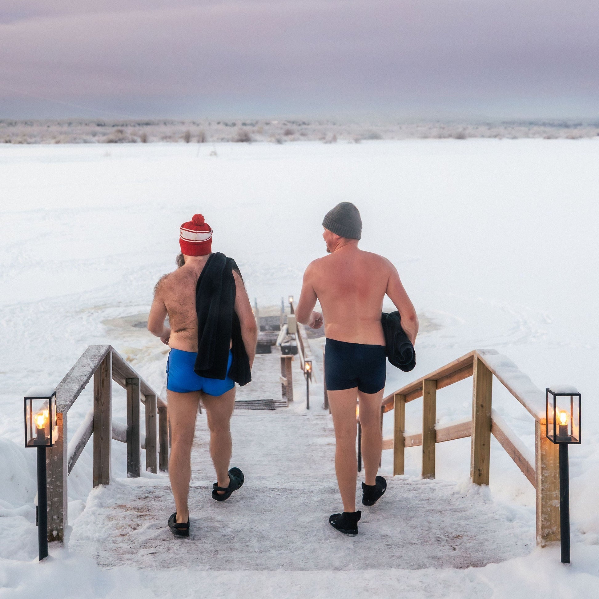Two people in swimsuits walking up a snowy staircase with candles on a cold day.