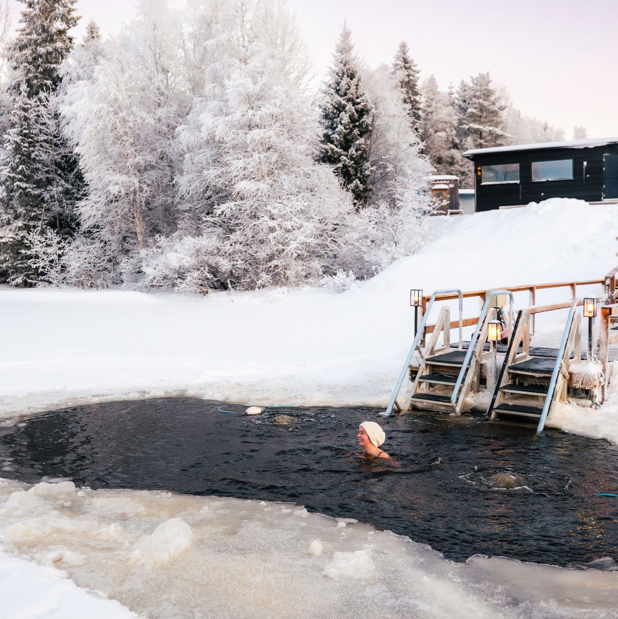Person swimming in a frozen lake with snow-covered trees and a building in the background