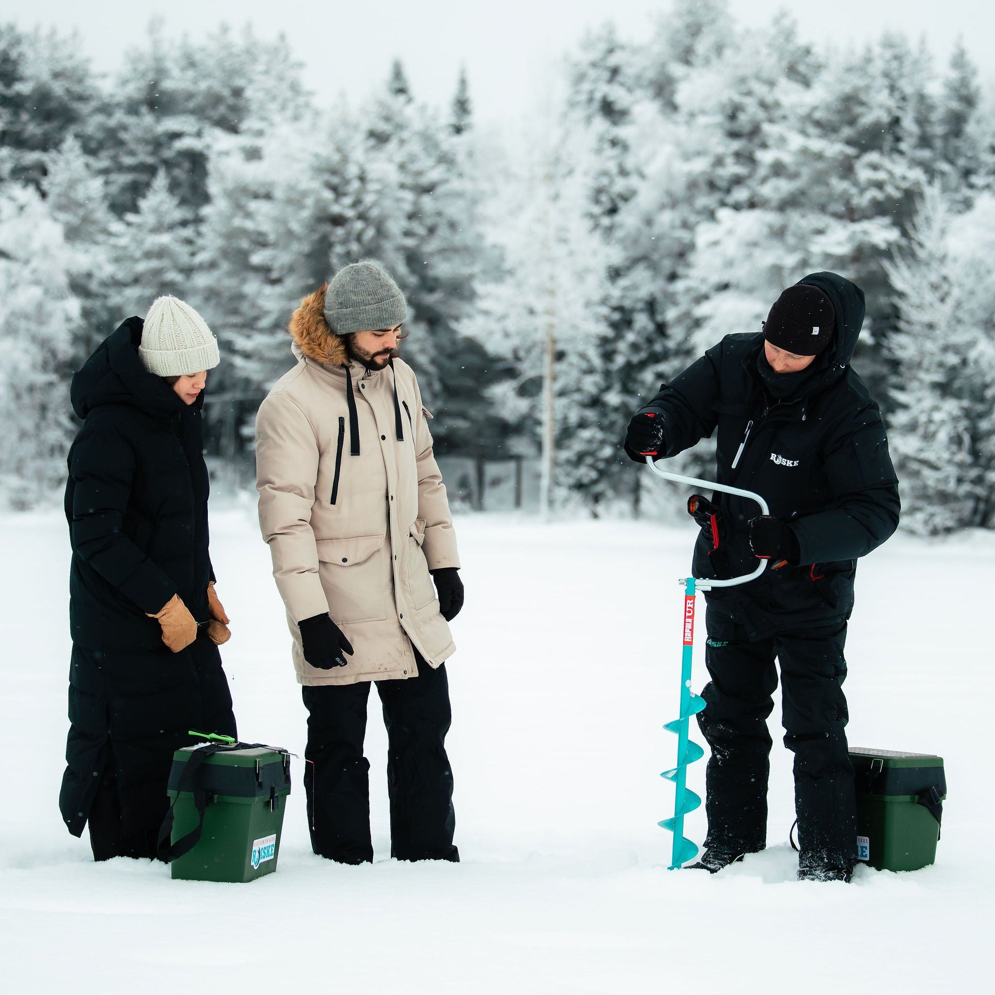 Three people in winter clothing in a snowy landscape with one using an ice auger.